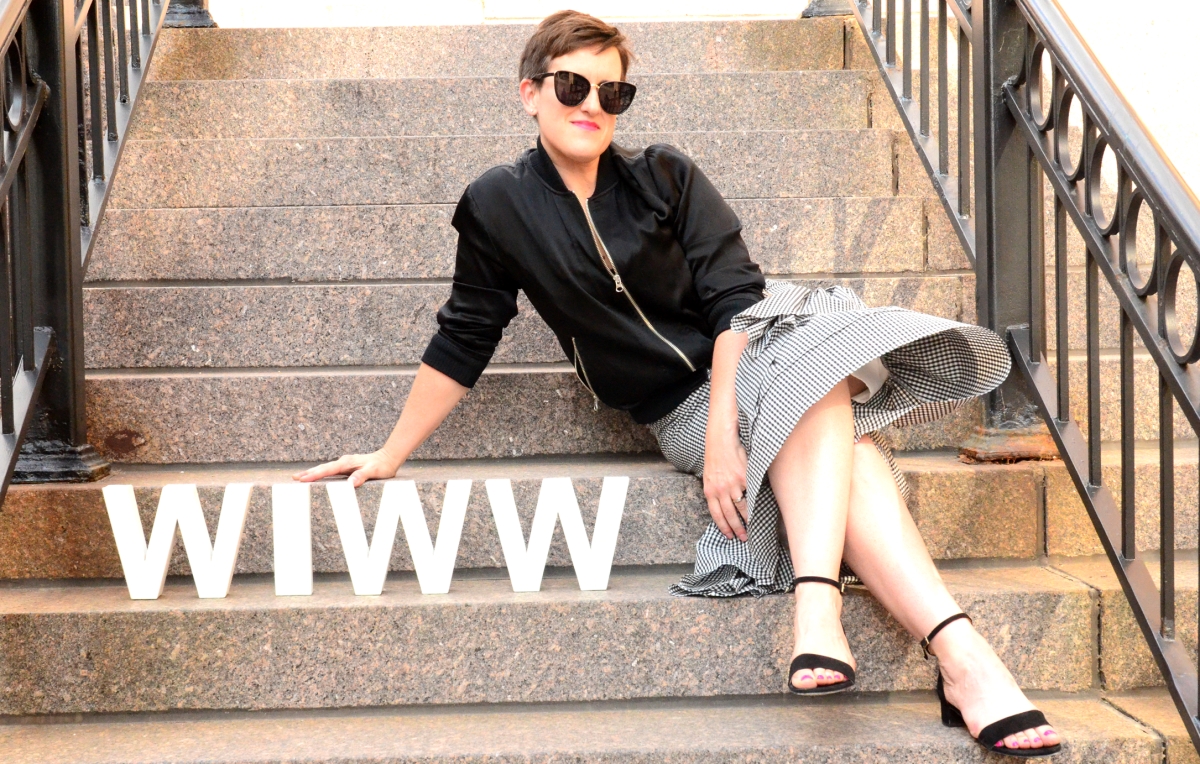Woman sitting on steps with gingham skirt, bomber jacket and sunglasses